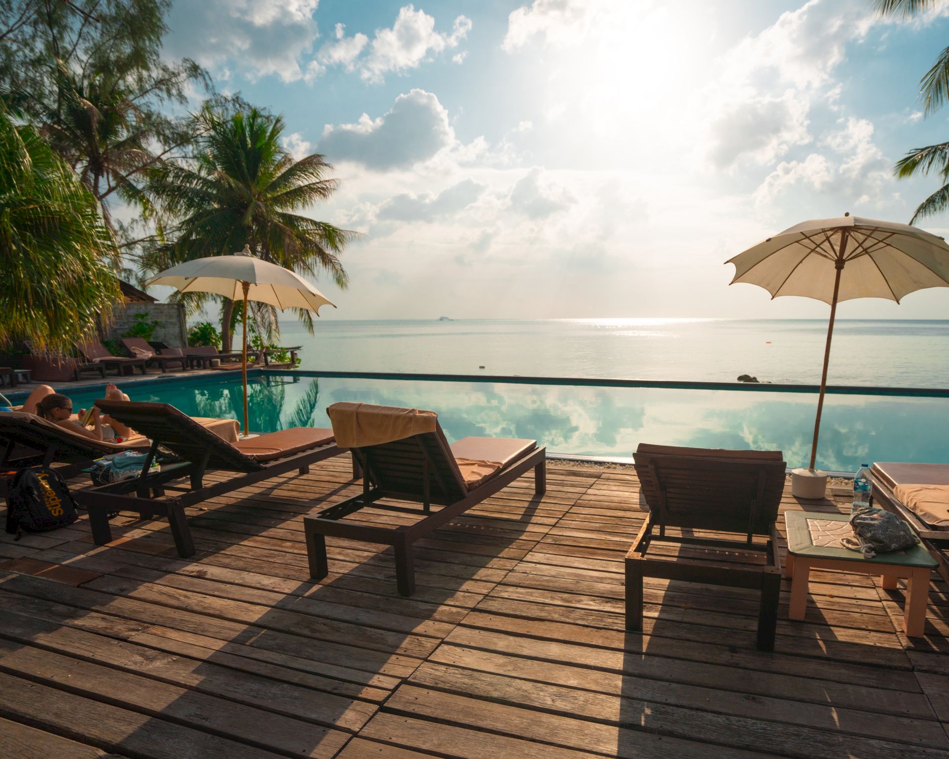 A tropical seaside resort deck with pool, lounge chairs under umbrellas, palm trees, and a bright ocean horizon under clouds.