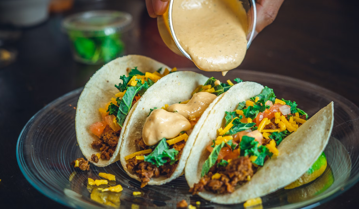 Three tacos on a plate with cilantro, shredded cheese/corn, and sauce being poured on top.