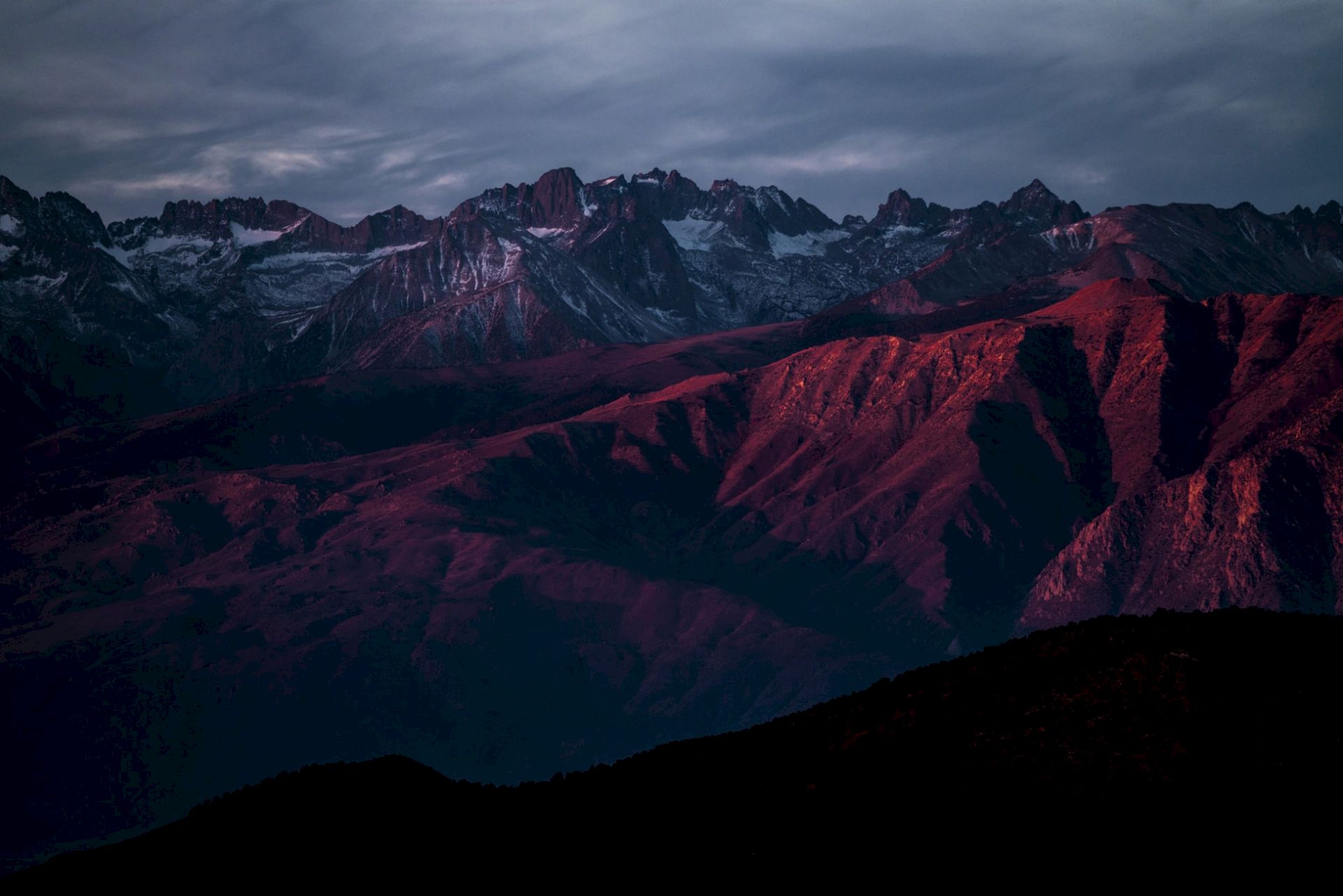 Dramatic mountain range at twilight, jagged peaks and snowfields glowing red under stormy clouds.