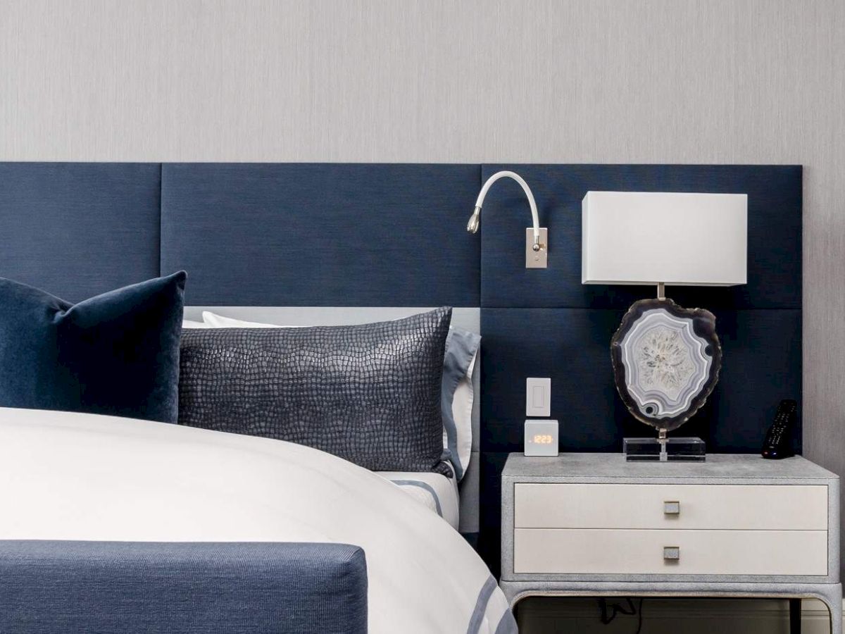 The image shows a modern bedroom with a blue headboard, stylish pillows, a bedside table with a lamp, and a wall light fixture.