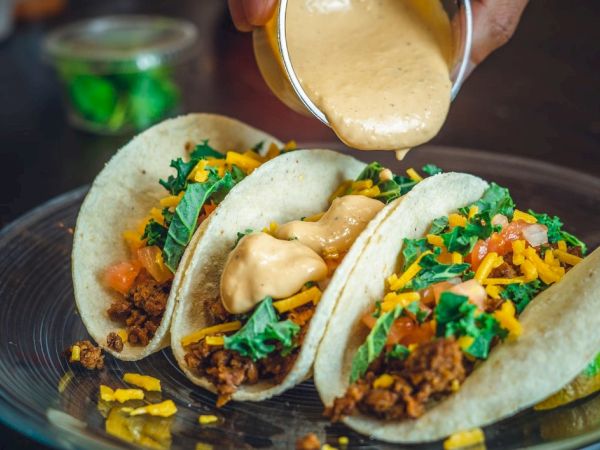Three tacos filled with ground meat, shredded cheese, tomatoes, and greens are being topped with a creamy sauce from a small container.