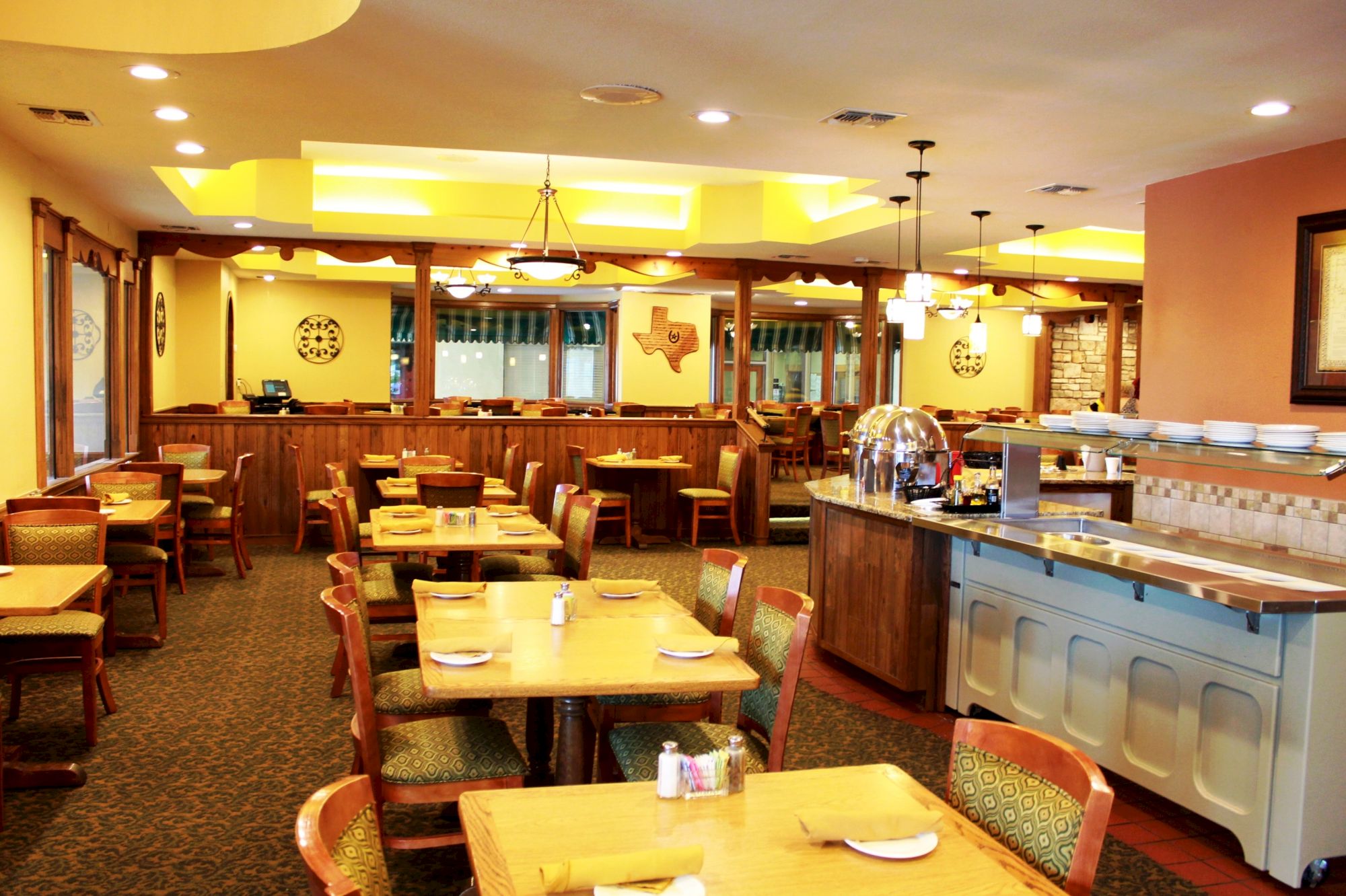 The image shows an empty restaurant with wooden tables and chairs, warm lighting, and a buffet counter, creating a cozy dining atmosphere.