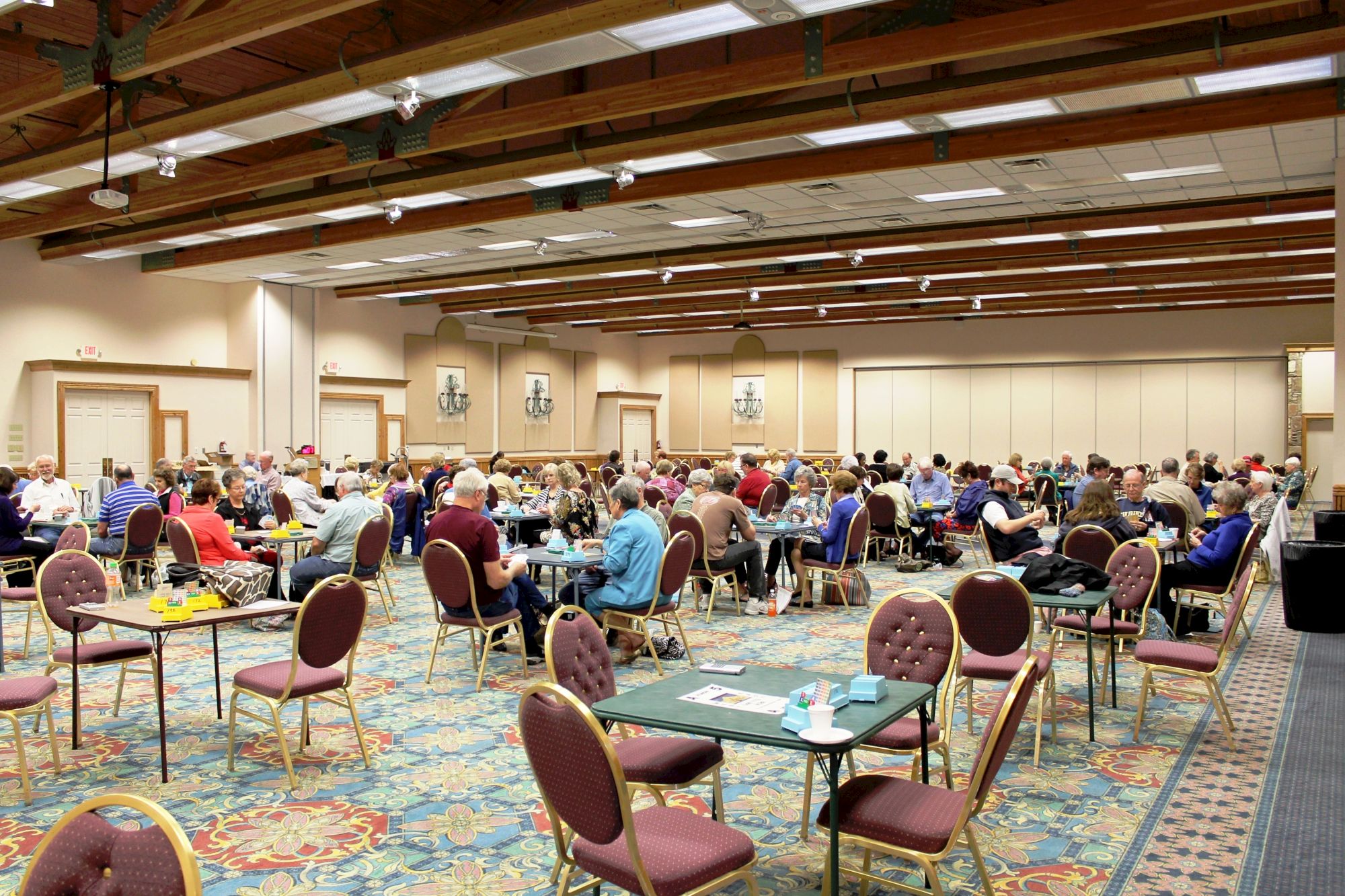A large banquet hall filled with many round tables and people dining, with chandeliers and a patterned carpet, chairs around tables.