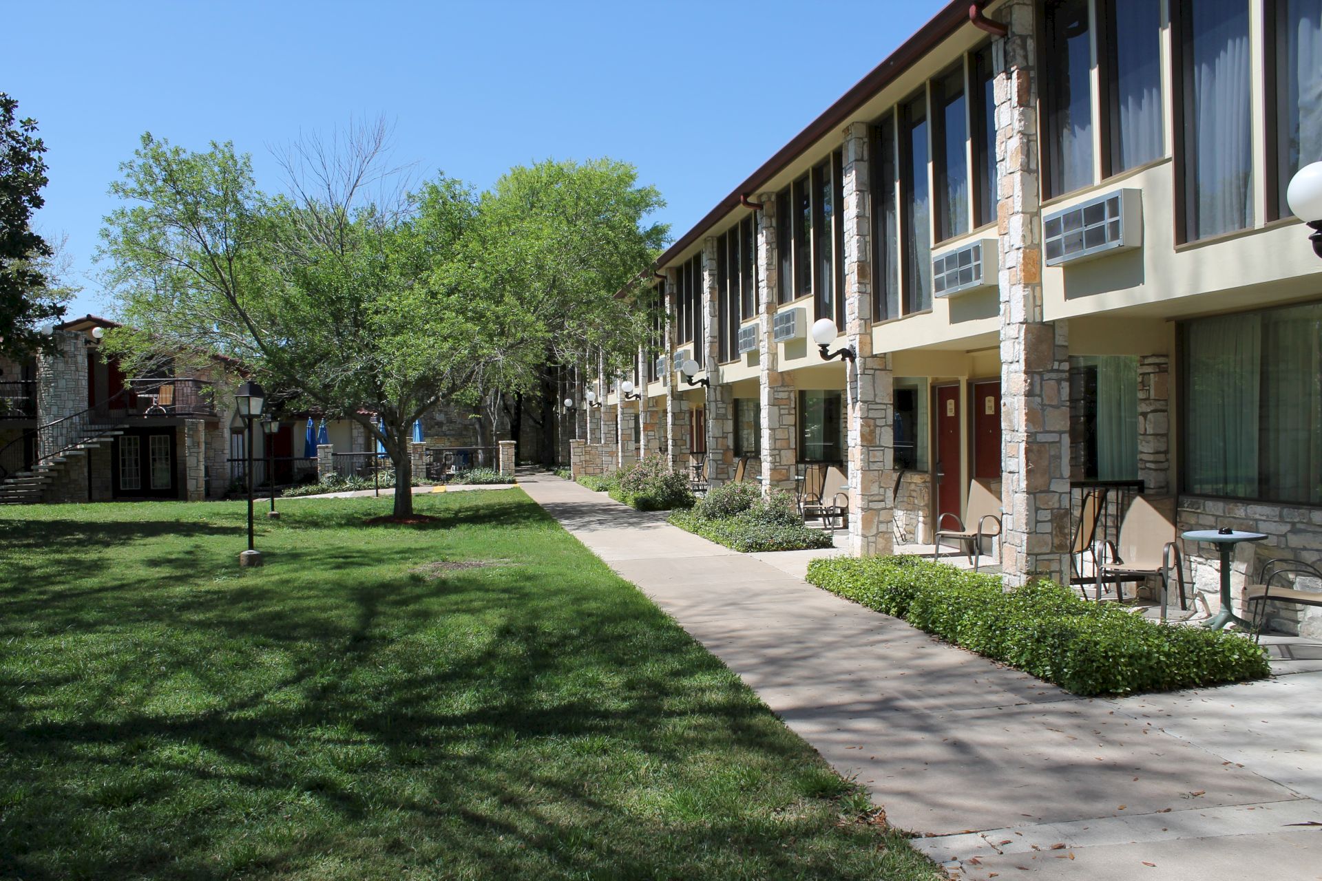 Sunlit courtyard beside a stone-and-brick hotel building with windows, a walkway, green lawn, trees, and outdoor tables.