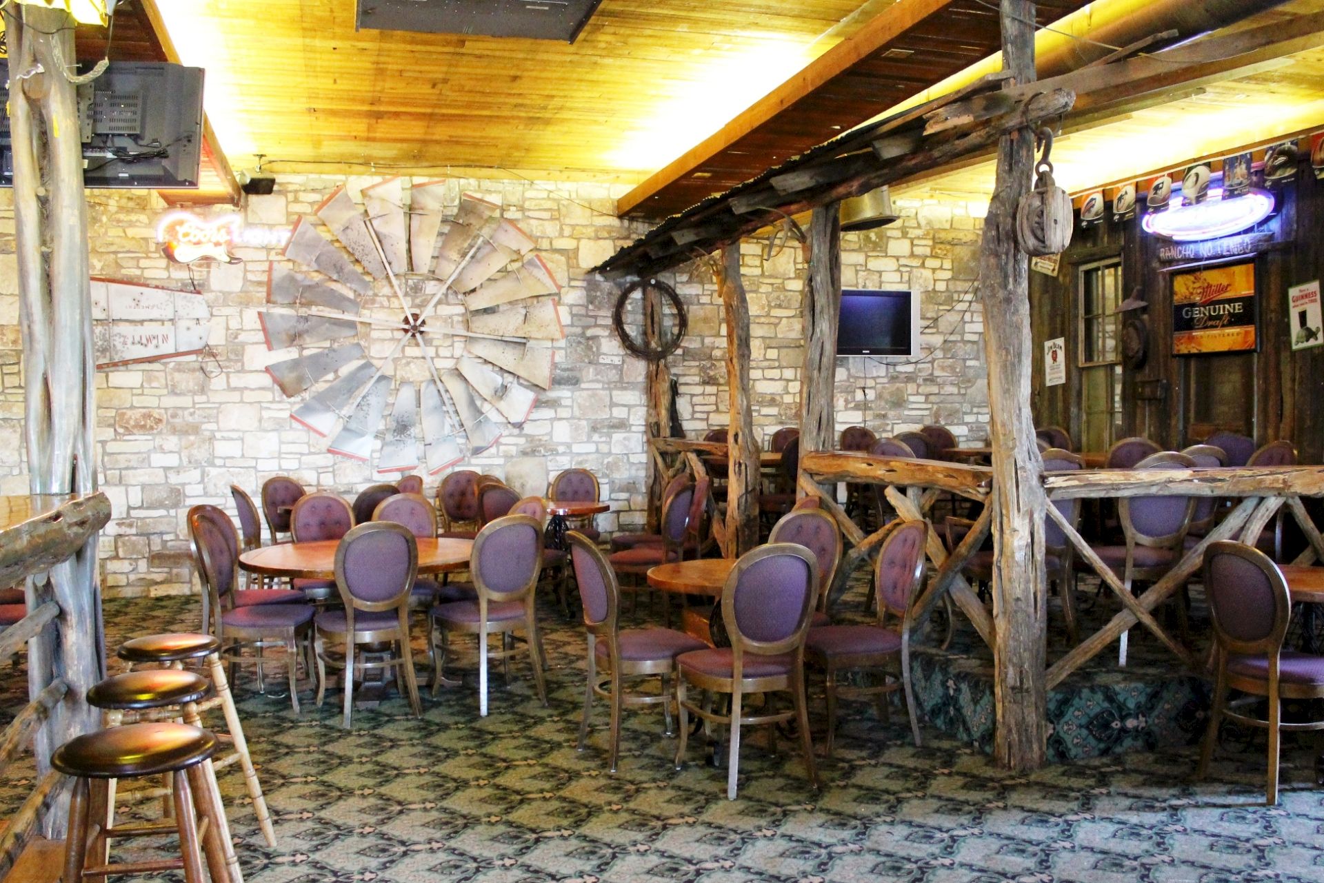 A rustic bar interior with purple chairs and tables, exposed beams, stone walls, and a large metal waterwheel decoration behind it.