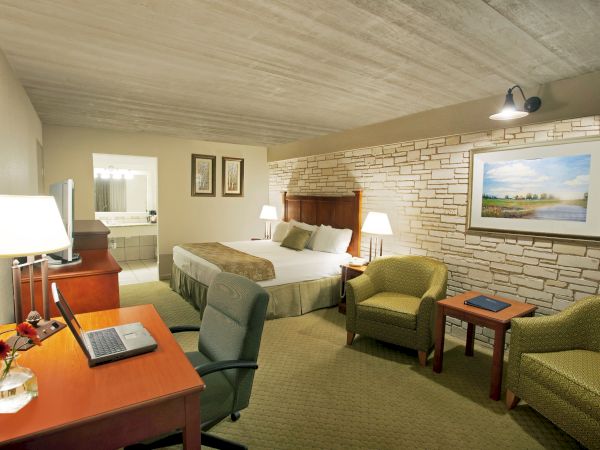 The image shows a cozy hotel room with a bed, desk, chairs, and lamps. There's a brick wall and a painting for decoration.
