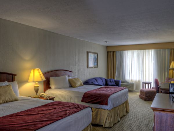 The image shows a hotel room with two beds, lamps, a sofa, a window with curtains, and a TV on a stand.