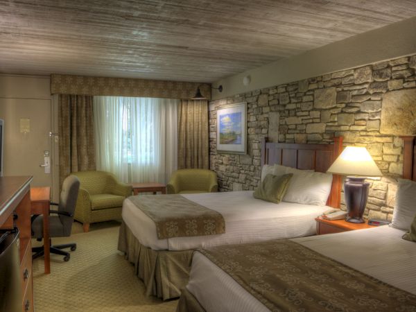 A cozy hotel room with two beds, a stone accent wall, a lamp, desk, chair, and window with curtains, creating a warm, inviting atmosphere.