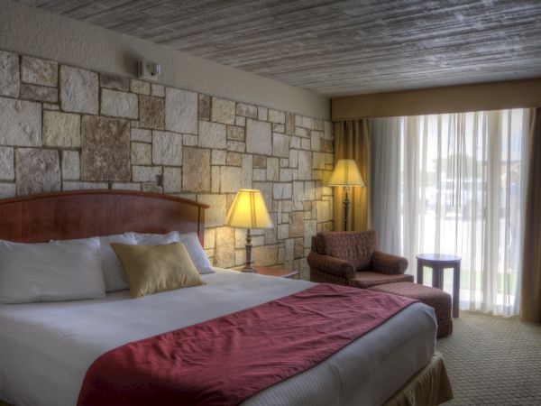 A cozy hotel room with a bed, two lamps, an armchair, and a window with curtains.