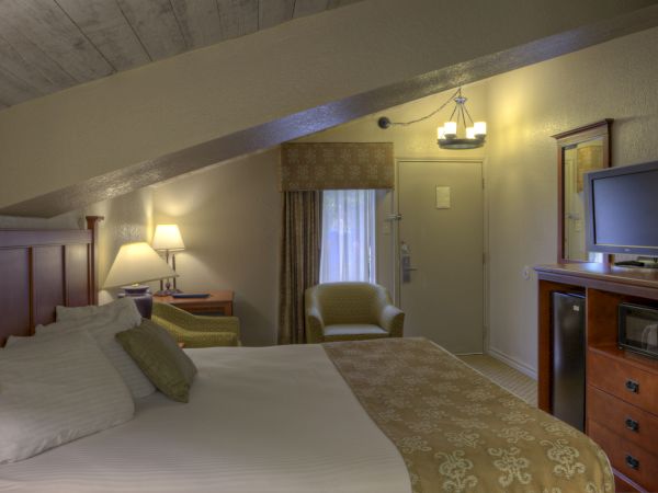 A cozy hotel room features a slanted ceiling, bed, TV, chair, and small refrigerator, with warm lighting and neutral tones throughout.