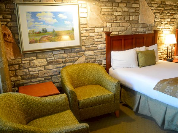 A cozy hotel room with a stone wall, bed, two yellow armchairs, a side table, a lamp, and a landscape painting above the bed.