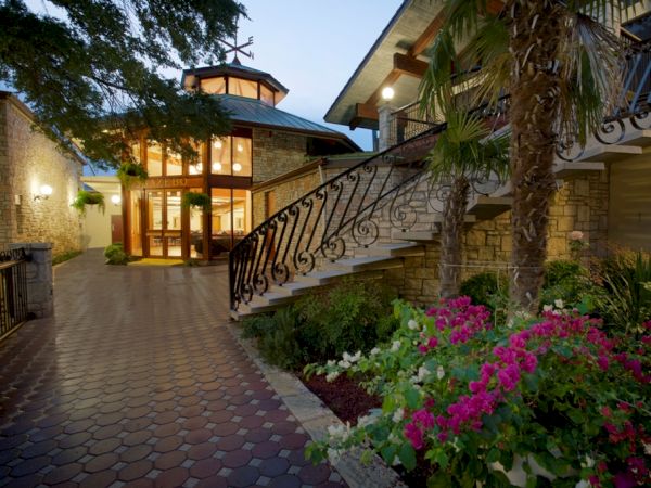 A beautifully lit entrance with a curved staircase, vibrant flowers, and lush greenery, leading to a modern building with large windows.