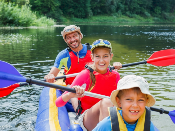 A family is enjoying a canoe ride on a calm river, paddling together in summer attire with life vests, surrounded by lush green trees.