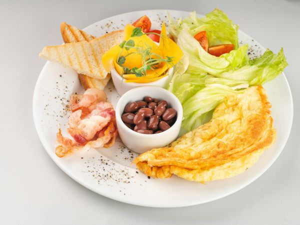 A breakfast plate with toast, bacon, beans, an omelet, lettuce, and fruit in a bowl, garnished with herbs.