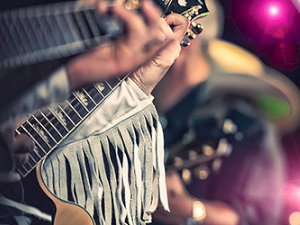 A close-up of musicians playing guitars, with one wearing a fringed jacket. Bright stage lights create a lively atmosphere.