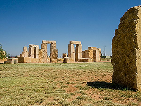 The image shows a sculpture resembling Stonehenge, set in a grassy area with a clear blue sky in the background.