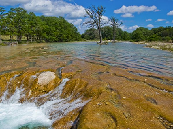 A scenic river landscape with smooth flowing water, rocks, lush green trees, and a blue sky with clouds in the background.