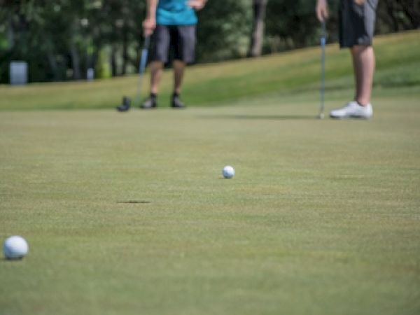Two individuals on a putting green, focusing on two golf balls, with one person about to putt in the foreground.