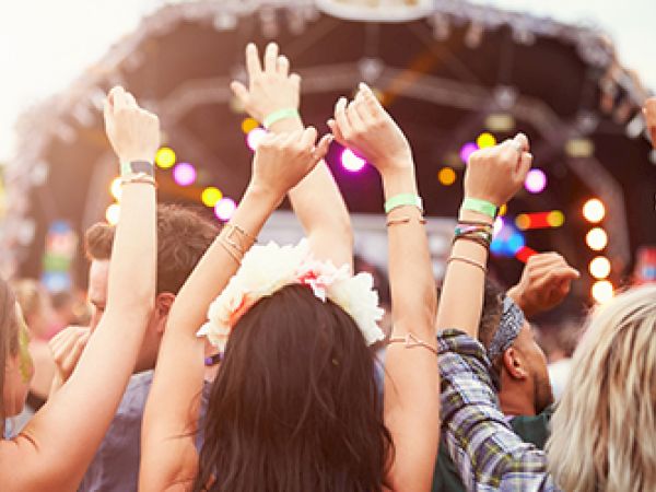People are enjoying a music festival, with arms raised, vibrant lights, and a festive atmosphere in front of a stage.