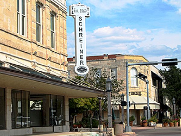 A street scene with historic buildings, featuring a sign that reads "Schreiner" on one of the structures, under a partly cloudy sky.
