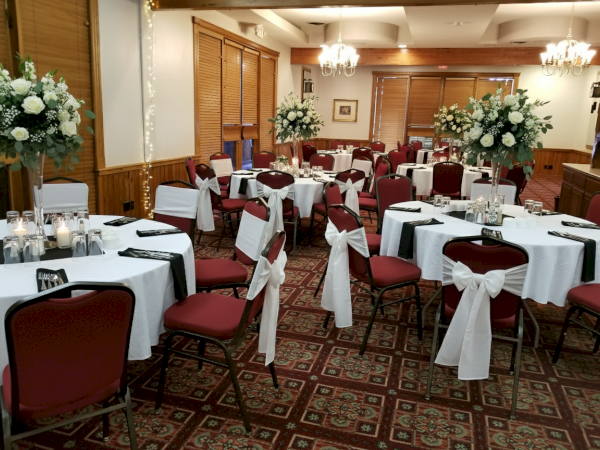 The image shows an elegantly decorated banquet hall with round tables, white tablecloths, red chairs, and floral centerpieces arranged for an event.