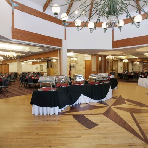 A spacious banquet hall with a buffet setup, decorated with chandeliers and wooden accents.