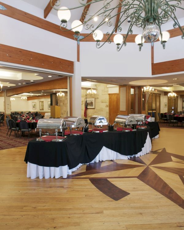 A spacious banquet hall with a buffet setup, decorated with chandeliers and wooden accents.