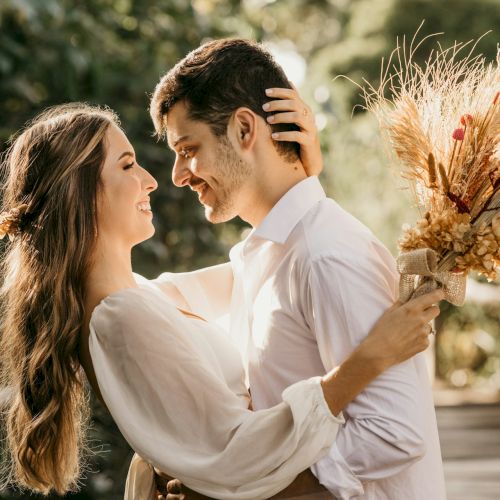 A couple embraces lovingly outdoors; the woman holds a bouquet of dried flowers. They both appear happy and are dressed in light clothing.
