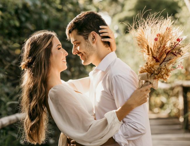 A couple embraces lovingly outdoors; the woman holds a bouquet of dried flowers. They both appear happy and are dressed in light clothing.