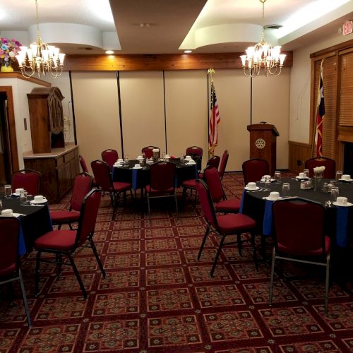 The image shows a banquet room with round tables set with tableware, red chairs, chandeliers, and flags near a lectern.