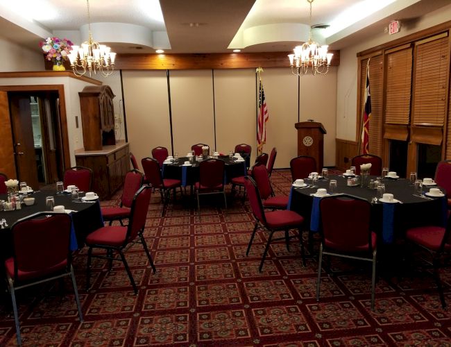 The image shows a banquet room with round tables set with tableware, red chairs, chandeliers, and flags near a lectern.