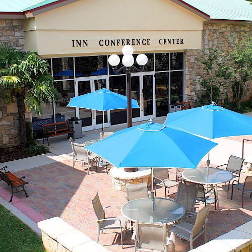 An outdoor patio with round tables, blue umbrellas, and chairs in front of a stone building labeled “Inn Conference Center.”