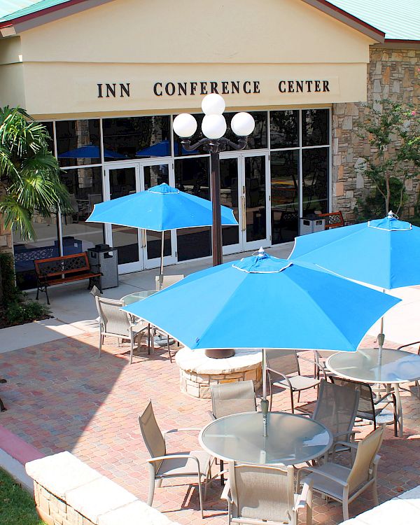 An outdoor patio with round tables, blue umbrellas, and chairs in front of a stone building labeled “Inn Conference Center.”
