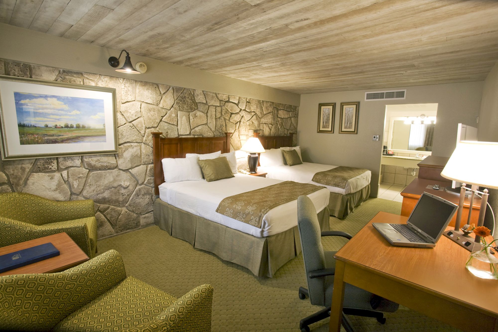 The image shows a hotel room with two beds, a desk with a laptop, two chairs, and a bathroom in the background.