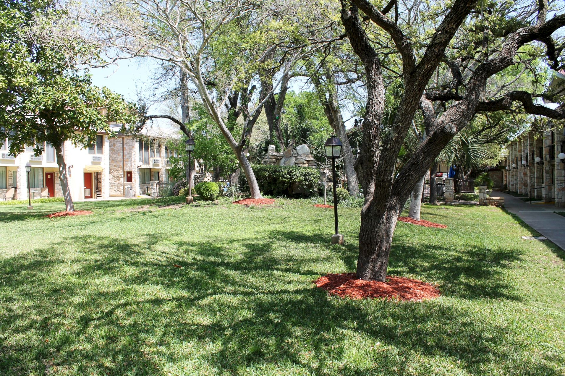A landscaped courtyard garden with large trees, green lawn, shrubs, and stone-and-brick buildings lining a walkway.