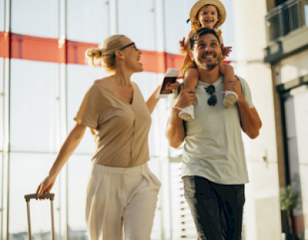 A family happily walks through an airport, with a woman pulling luggage and a man carrying a child on his shoulders.