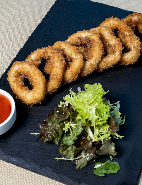 Fried calamari rings with a side of red dipping sauce and mixed greens, served on a dark rectangular slate.
