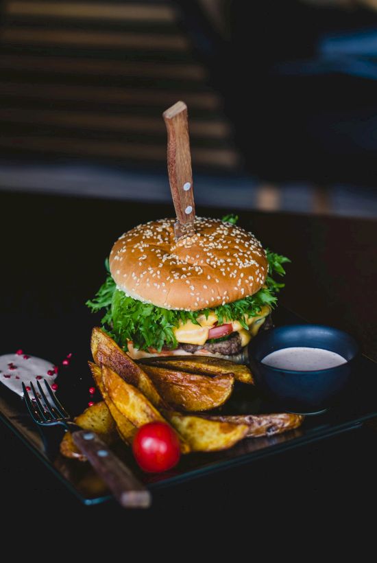 A burger with a knife through it, accompanied by potato wedges, a cherry tomato, and sauce, served on a black plate.