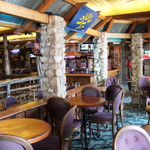 The image shows an empty rustic bar with wooden tables, stone pillars, and a few televisions. The setting has a cozy, lodge-like atmosphere.
