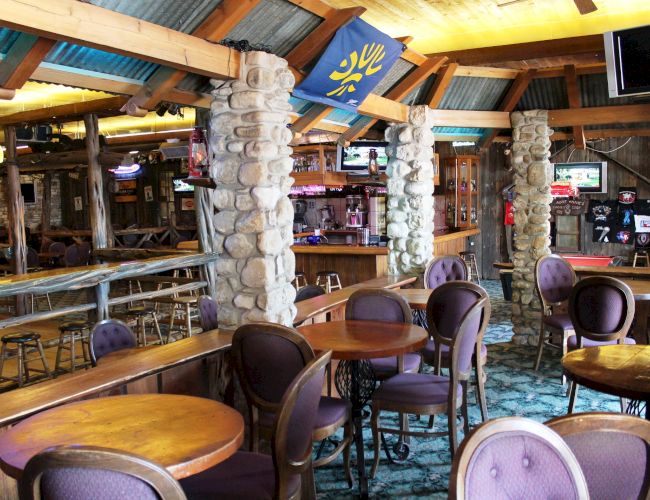 The image shows an empty rustic bar with wooden tables, stone pillars, and a few televisions. The setting has a cozy, lodge-like atmosphere.