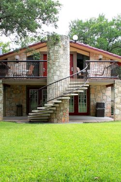 A two-story stone house with a wide lawn, balcony, exterior stairs, trees, and blue patio umbrellas in a sunny yard.