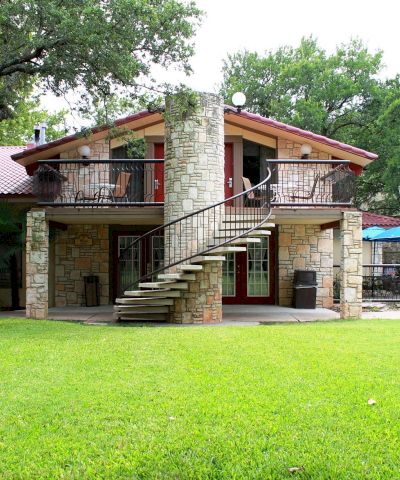 A two-story stone house with a wide lawn, balcony, exterior stairs, trees, and blue patio umbrellas in a sunny yard.