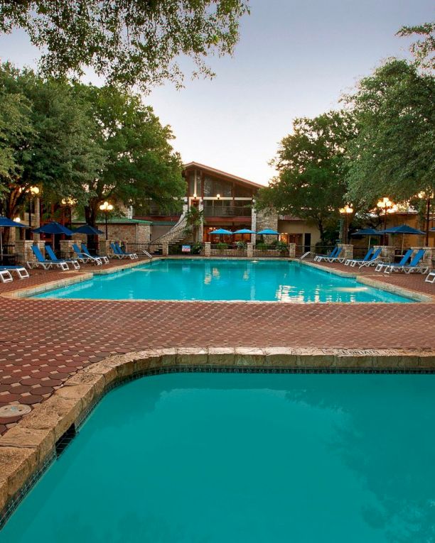 A resort pool area with two turquoise pools, lounge chairs, umbrellas, trees, and a central building, all bathed in late-day light.