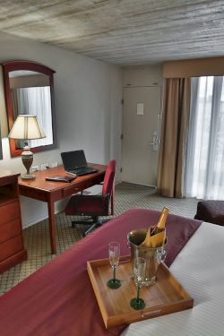 A hotel room with a bed, desk and chair, a TV on a wooden dresser, a sitting area with sofa and lamps, and a tray with drinks on the bed.
