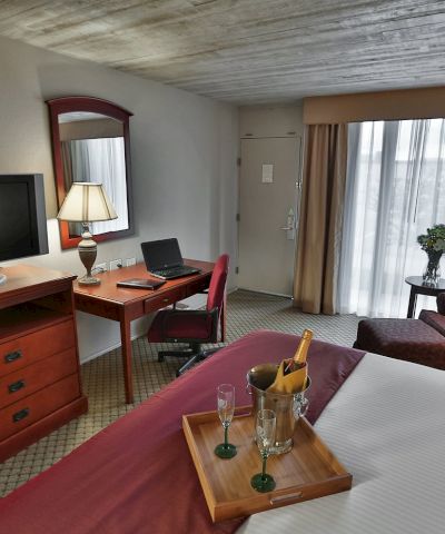 A hotel room with a bed, desk and chair, a TV on a wooden dresser, a sitting area with sofa and lamps, and a tray with drinks on the bed.