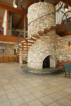 A spacious hotel lobby with a stone spiral staircase, curved stone fireplace, seating area, piano, and warm wooden beams.