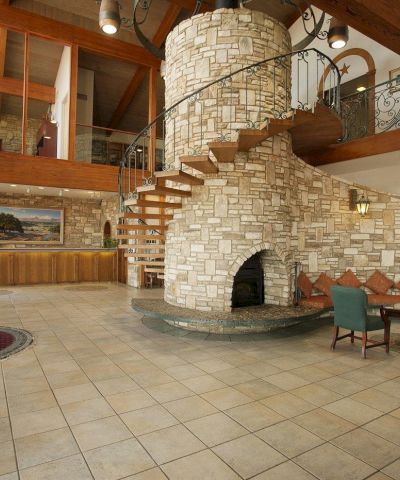 A spacious hotel lobby with a stone spiral staircase, curved stone fireplace, seating area, piano, and warm wooden beams.