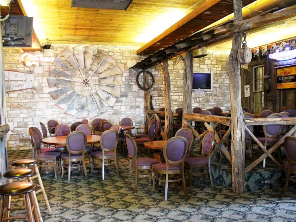 A rustic restaurant interior with wooden beams, round tables, purple chairs, and decorative elements on brick walls, including a TV.