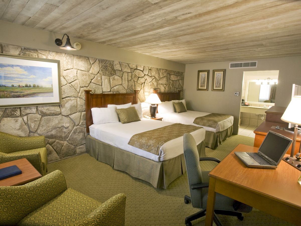 The image shows a hotel room with two beds, a stone accent wall, a desk with a laptop, and comfortable seating.
