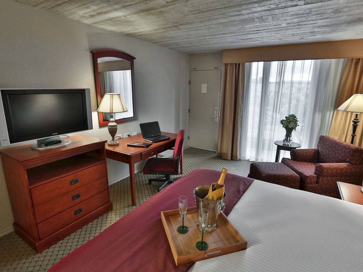 The image shows a hotel room with a bed, tray, TV, desk, chair, and a small seating area with a couch and lamp near a window.